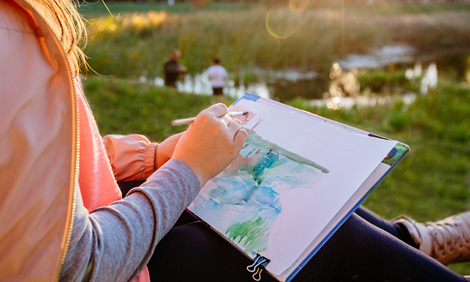 Girl water colour painting outside overlooking a lake on a summer evening.